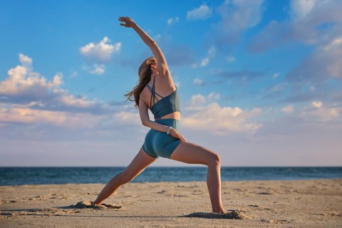 On beach sand, a woman holds a half-bound Viparita Virabhadrasana yoga variation, Reverse Warrior,
