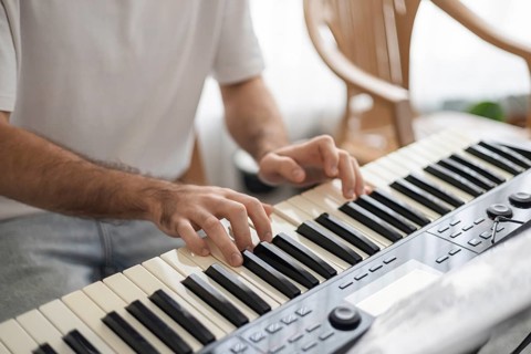A young man with dark hair plays a keyboard at home. The setting is cozy, with wooden chairs and