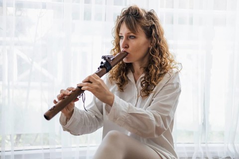 A woman plays a wooden flute by a window, creating a serene musical atmosphere