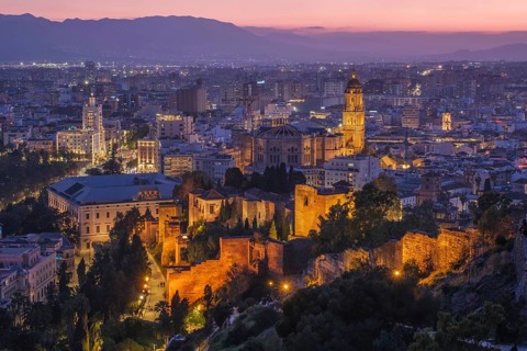 Illuminated Malaga Cathedral dominates the dusk skyline in Spain, rising behind golden Alcazaba