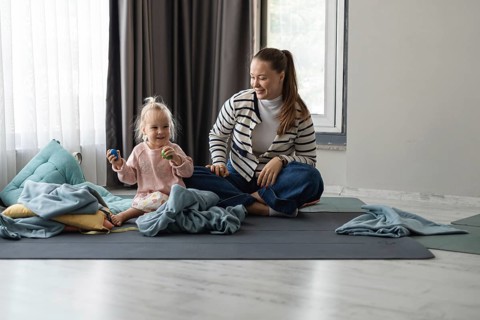 A young Caucasian woman with long brown hair sits on a mat with a toddler girl. The girl has blonde
