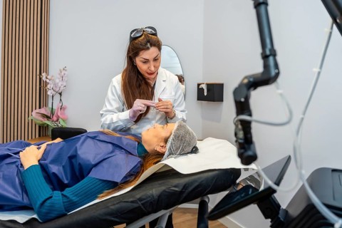 Aesthetic specialist in a white lab coat and gloves preparing a woman on a treatment bed for a