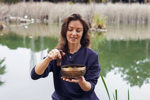 A young Caucasian woman with brown hair sits by a serene lake, holding a wooden bowl filled with