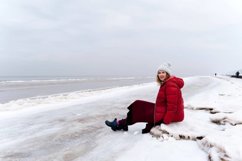 A woman in warm winter clothes rests sitting on the beach, A woman in a red coat sits on a snowy