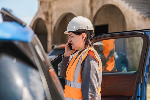 Worker woman in a hard hat and safety vest talks on her phone near a blue car. The scene is set