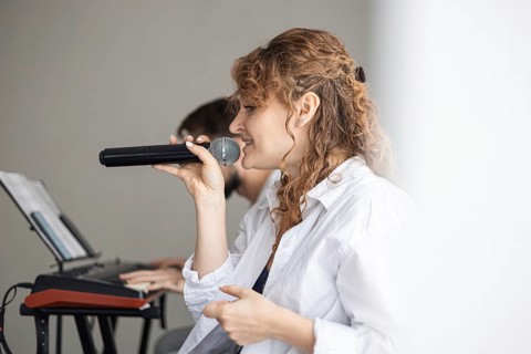 A young Caucasian woman with curly hair sings into a microphone. A man plays a keyboard in the