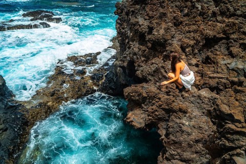 Woman crouching on rugged volcanic rock formations by the ocean, watching turquoise waves crashing