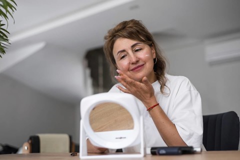 A woman smiles as she applies a skincare product to her face in a well-lit indoor space. The