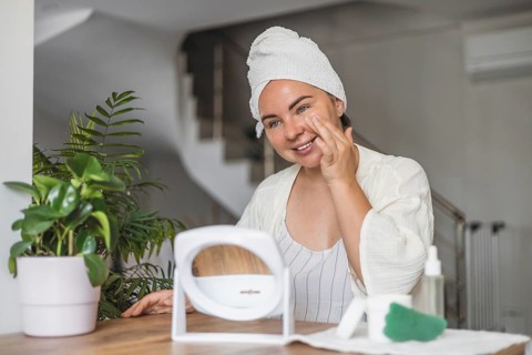 A young Caucasian woman with brown hair in a towel smiles while applying gua sha on her face. A