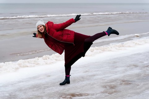 Cheerful woman on the beach in the arabesque pose in winter, A woman in a red coat and winter hat