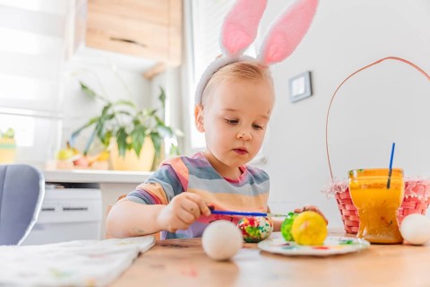 Toddler girl wearing pink bunny ears sits at table in kitchen decorated for Easter. Colorful eggs