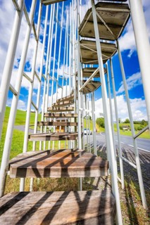 Outdoor wooden and metal staircase leading up to the sky away from a road surrounded by grasses,