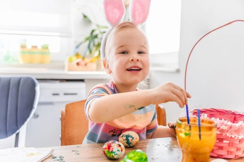 A child wearing bunny ears sits at a table decorated for Easter. Colorful eggs are placed on the
