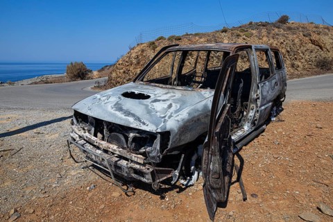 A burnt out car stands on the side of the road in front of a dry coastal landscape, forest fire,