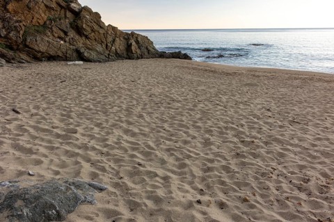 Beach-level viewpoint over an empty sandy cove with rippled sand, a rocky cliff, calm sea, and