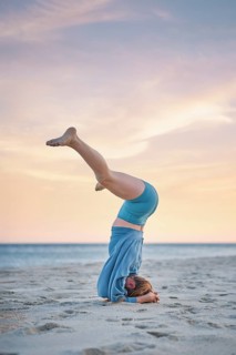 On beach sand, a woman holds Salamba Sirsasana Konasana yoga, a Wide-Legged Headstand, with her