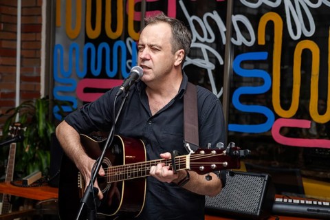 A middle-aged Caucasian man with short hair sings into a microphone while playing an acoustic