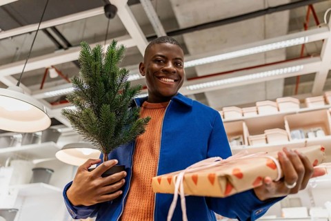 A person stands in a store holding a small potted tree in one hand and a wrapped gift in the other.