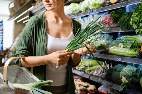 Woman holds green onions while exploring vegetable section in grocery store. She wears white top