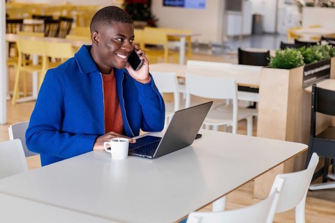 A young African man with short hair is sitting at a table in a modern cafe. He is using a laptop