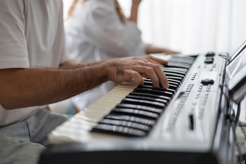 A close-up of a man's hand playing a keyboard in a cozy home setting. A woman is seated nearby,