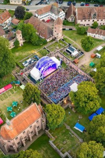 Outdoor concert in a historic setting with numerous spectators, surrounded by trees and old