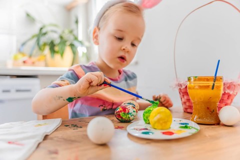 Toddler girl in dress wearing bunny ears painting Easter egg with brush, basket for eggs is near