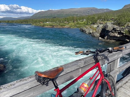 Red bicycle standing on a wooden bridge overlooking a rushing river and mountains, Sjoa,
