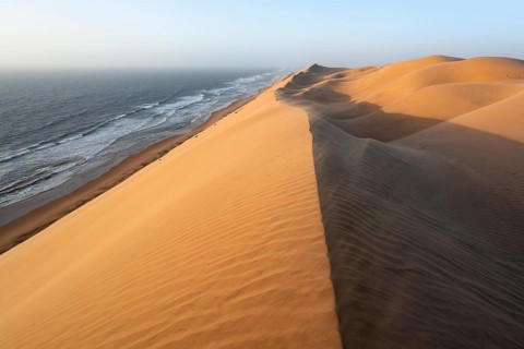 Sand dunes with wave patterns and sea in evening light, wind blowing sand across the dunes,