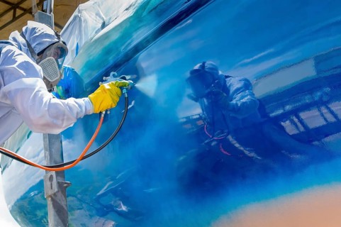 Worker spray paints the hull of a sailing yacht during professional marine maintenance, wearing