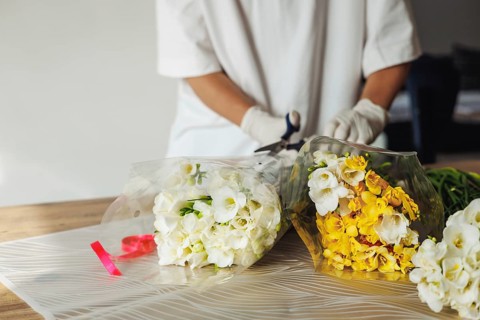 A florist, wearing gloves, is carefully arranging white and yellow flowers in wrapping paper.