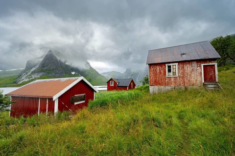 Red wooden houses in green mountain landscape with dramatic clouds, Vindstad, Reinefjord, Lofoten,
