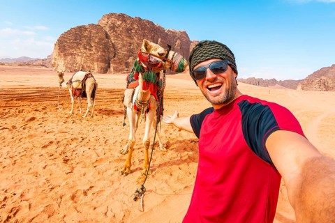 Smiling man traveler taking selfie standing near saddled camel orange sand valley sandstone rock