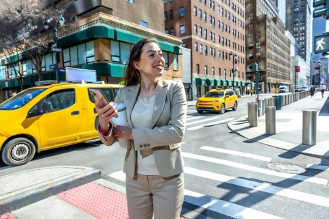 Young businesswoman standing smiling outdoors, holding a mobile phone and coffee while crossing a