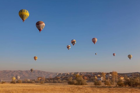 Several hot air balloons soar into the bright blue morning sky over an idyllic village and vast