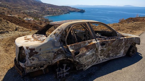 A rusty, burnt car with a view of the blue sea and rocky hills, forest fire, fire damage, fire on
