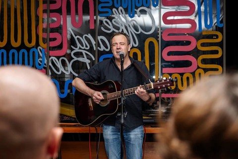 A middle-aged Caucasian man with short brown hair sings into a microphone while playing an acoustic