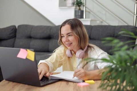 Young blond European woman with headphones lying on floor near laptop and notebook in modern living