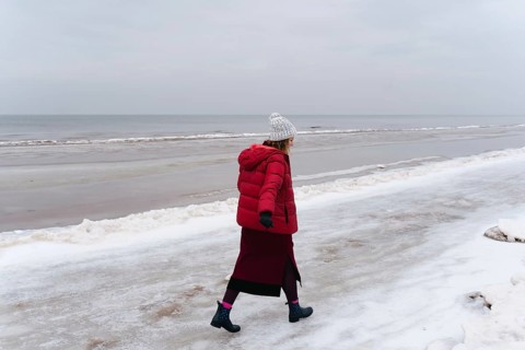 A woman in warm clothes walks along the sea in winter, A woman in a red coat walks alone on a snowy