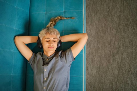 Woman lies on blue couch with headphones on, eyes closed, and hands resting behind head. Soft light