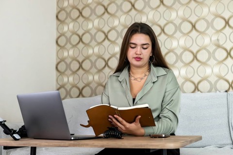 A young Hispanic woman with long brown hair reads a notebook while sitting at a desk with a laptop.