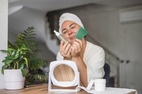 A young Hispanic woman with a towel on her head holds a gua sha tool and a serum bottle. A mirror