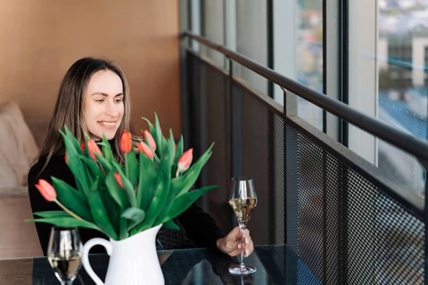 A young woman sits at a table by the window and holds a glass of champagne. There is a vase of
