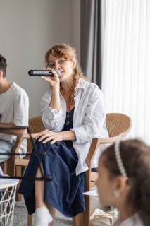 A young Caucasian woman with curly hair sings into a microphone at a home concert. Two children