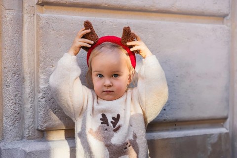 A young Caucasian girl with blonde hair wears a festive sweater and reindeer antlers. She poses