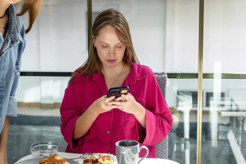 Woman sits at a table, looking at her phone. She has a cup next to her and some food on a plate.
