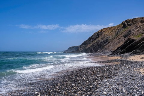 Pebble beach with cliffs in the background, waves gently hitting the coast under a clear blue sky,
