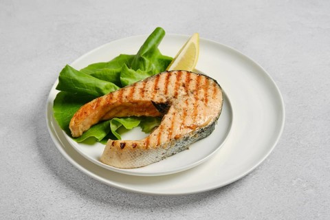 Grilled salmon steak on white plate next to fresh salad leaves and slice of lemon. The food is