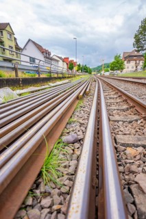 Railway tracks with gravel flanked by buildings and vegetation under cloudy skies, Leonhard Weiß