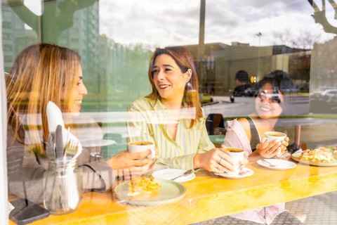 Three smiling women friends sitting at a wooden cafe table, enjoying coffee, conversation and